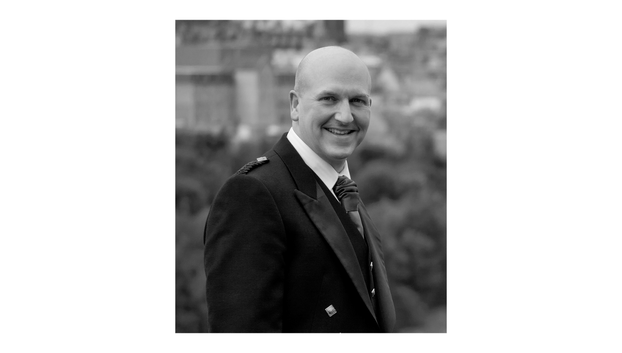 A side profile of Stevie smiling gently in monochrome, with Edinburgh rooftops in the distance and the Declaration tartan sash in vivid colour. A candid, warm portrait of The Tartan Artisan.