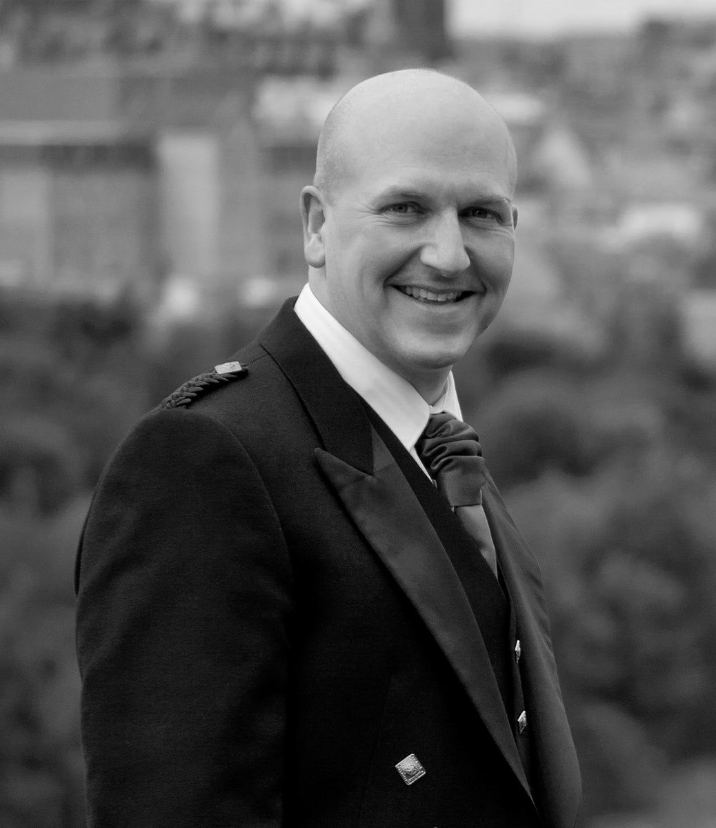 A side profile of Stevie smiling gently in monochrome, with Edinburgh rooftops in the distance and the Declaration tartan sash in vivid colour. A candid, warm portrait of The Tartan Artisan.